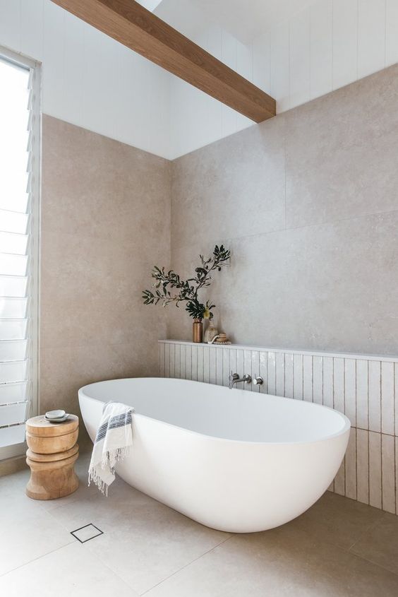 a neutral coastal bathroom with neutral large scale tiles and white skinny ones, an oval tub, a wooden beam and a wooden stool