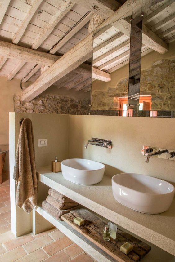 a neutral farmhouse bathroom with stone walls, with a whitewashed ceiling with beams, a double built-in vanity and mirrors