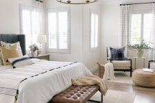 a neutral farmhouse bedroom with a grey bed, a brown leather bench, neutral chairs, a jute pouf and a chandelier