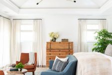 a neutral farmhouse bedroom with a large bed, a blue loveseat, rust chairs, wooden stools and a dresser, potted plants and neutral textiles