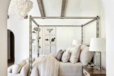 a neutral refined bedroom with wooden beams, a metal canopy bed, neutral bedding, a white sofa and a cool rug plus a lovely chandelier