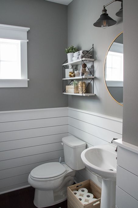 a pretty modern farmhouse bathroom with grey walls and white planks on the walls, a round mirror, a pedestal sink, a small yet practical pendant shelf