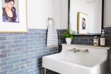 a pretty powder room with blue tiles on the walls, a console sink, a mirror in a black frame and a pendant lamp plus a bold artwork