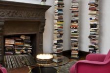a refined living room with a vintage fireplace with lot sof books and magazines, with book stacks, with bold pink chairs and a round glass table