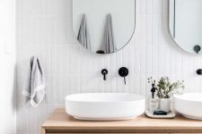 a refined modern bathroom clad with white skinny tiles, with a stone floor, a floating light-stained vanity, oval vessel sinks and oval mirrors