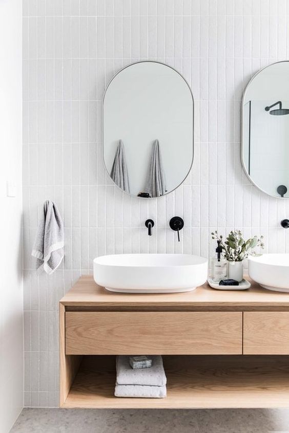 a refined modern bathroom clad with white skinny tiles, with a stone floor, a floating light-stained vanity, oval vessel sinks and oval mirrors