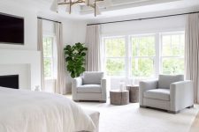a refined neutral bedroom with wooden beams, a fireplace, a bed and white chairs, potted plants and a chic chandelier