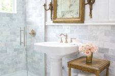 a refined vintage bathroom clad with marble subway tiles, a vintage pedestal sink, a vintage tiered table, a mirror in a gilded frame and vintage fixtures