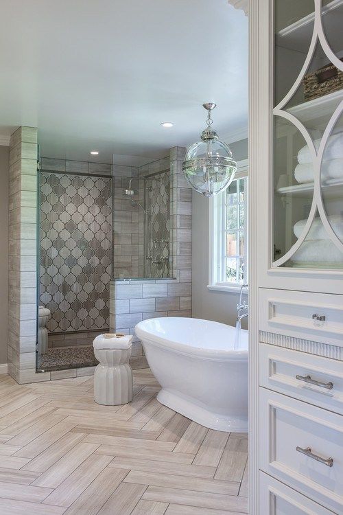a refined vintage glam bathroom clad with chervon tiles on the floor, arabesque ones in the shower space, a refined tub and a glass globe chandelier
