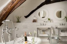 a refined white bathroom with white subway tiles, whitewashed floors, two free-standing sinks, round mirrors and dark stained beams