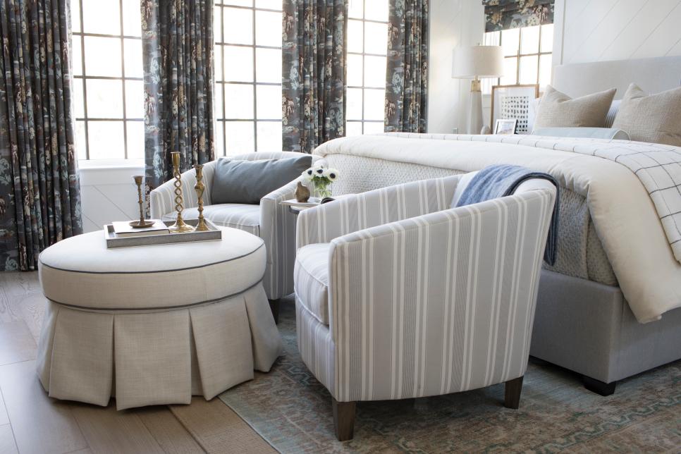 a retro bedroom with statement dark printed curtains, a neutral bed and striped chairs, a round ottoman and candlesticks