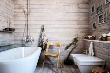 a rustic bathroom covered with weathered wood looking laminate, with wooden beams, a long open shelf and an oval tub plus pendant lamps