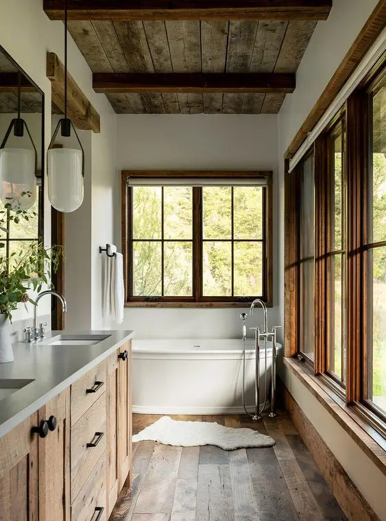 a rustic bathroom with a wooden floor, a ceiling with beams, a wooden vanity, a pendant lamp, some windows and a vintage bathtub