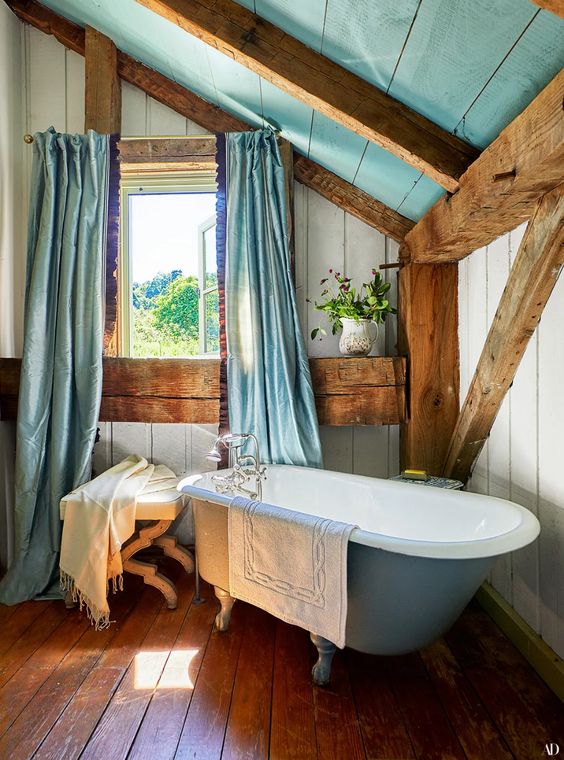 a rustic blue and white bathroom clad with wooden planks, a blue clawfoot tub, lots of wooden beams and pillars and blue textiles