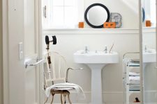 a shabby chic bathroom with neutral walls and grey planked floors, a pedestal sink, a round mirror, a vintage chair and a cart with glass shelves