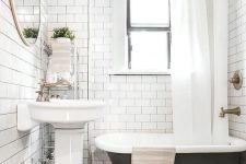 a small and cute bathroom with black and white tiles, white subway ones, a black clawfoot tub, a pedestal sink, a round mirror and a storage cart