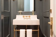 a small and elegant powder room with grey paneling, a console sink, chic sconces, white towels and touches of gold and brass