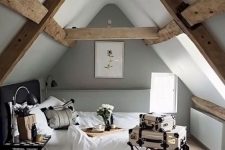 a small attic Scandi bedroom with wooden beams, a dark bed with neutral bedding, a folding nightstan and vintage suitcases for decor