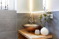 a small bathroom clad with concrete tiles, with wooden floors, a wooden vanity with a catchy and shiny vessel sink and a lit up mirror