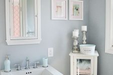 a small grey powder room with a pedestal sink, a small storage unit, a bright gallery wall and a small mirror cabinet plus a sconce