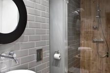 a small modern bathroom clad with grey and wood look tiles, a small shower space, a bowl sink, a round mirror and white appliances