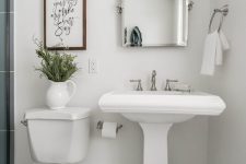 a small neutral bathroom with a pedestal sink, a mirror cabinet, a sconce, greenery, a sign and a shower space with blue tiles