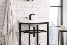 a stylish bathroom with faux white brick, a console sink, a round mirror in a frame, a black lantern and neutral textiles