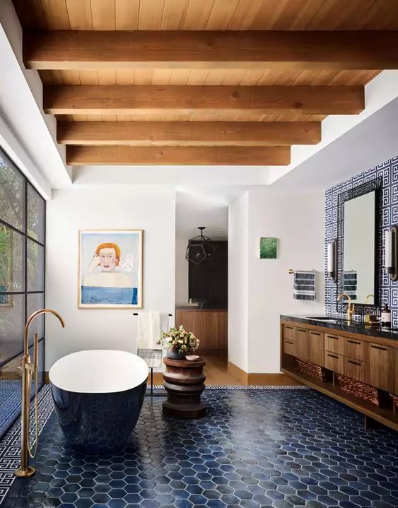 a stylish bathroom with wooden beams, navy hex tiles, a navy oval tub, a floating vanity and some art plus a glazed shower space