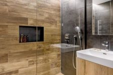 a stylish contemporary bathroom clad with black stone and wood look tiles, a niche, a light stained vanity and white appliances