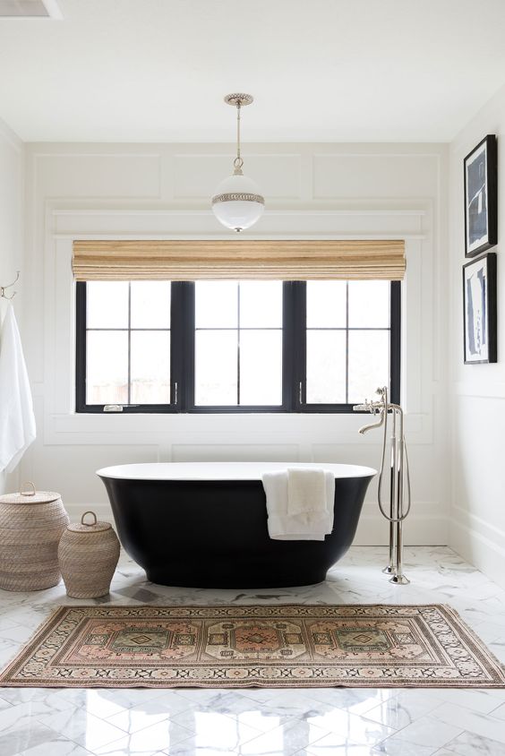 a stylish modern bathroom clad with white panels, with a black window frame, a black tub, a boho rug, a small gallery wall and baskets