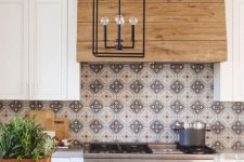a stylish white farmhouse kitchen with a wooden hood, a pretty patterned tile backsplash, a white and grey kitchen island and a black pendant lamp