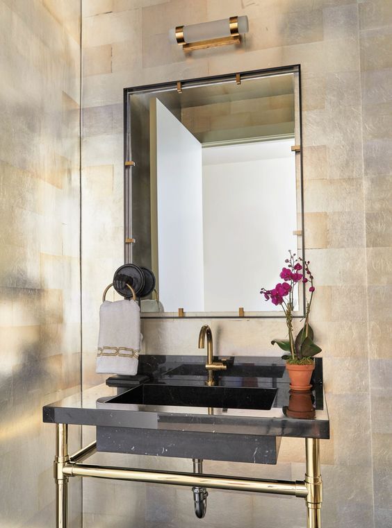 a super elegant powder room with metallic wallpaper walls, a black console sink, a mirror in a metal frame and a sconce over it