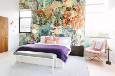 a unique bedroom with small windows, a statement colorful wall, a white bed and colorful bedding, a pink chair and a side table