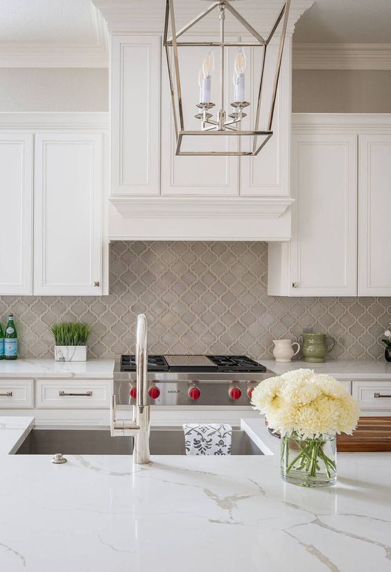 a vintage and refined white kitchen with shaker style cabinets, a grey arabesque tile backsplash, white stone countertops and chromatic appliances