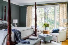 a vintage bedroom with grey paneled walls, a rich-stained cnaopy bed, white chairs, a blue ottoman and a view of the forest