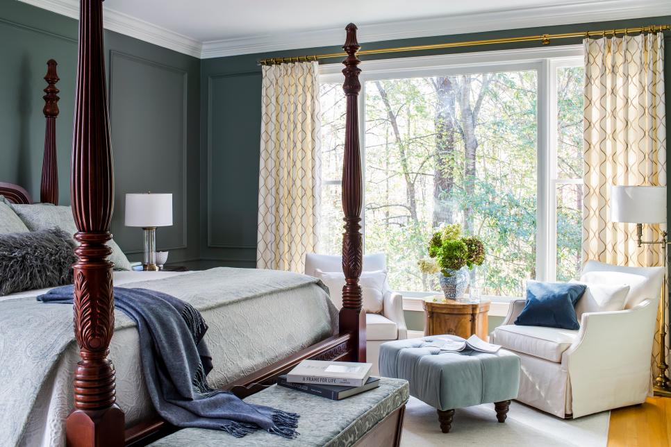 a vintage bedroom with grey paneled walls, a rich stained cnaopy bed, white chairs, a blue ottoman and a view of the forest