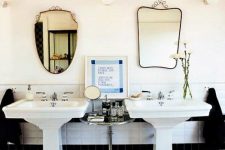 a vintage black and white bathroom with two pedestal sinks, black and white tiles, mismatching mirrors, a side table with various stuff