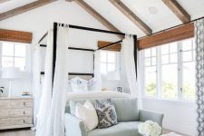 a vintage farmhouse bedroom with wooden beams, a canopy bed and a blue sofa, a large rug and printed curtains