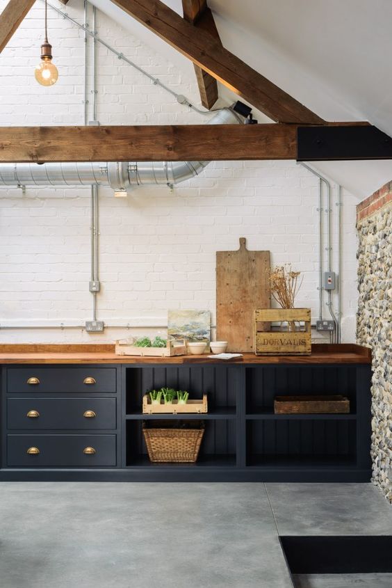 a vintage farmhouse kitchen with navy cabinets, wooden beams, a stone accent wall and rich-stained countertops and pendant lamps
