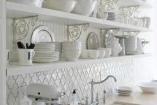 a vintage white kitchen with open shelves, a white arabesque tile backsplash, neutral stone countertops and silver fixtures is very refined