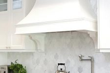 a vintage white kitchen with shaker cabinets, a large vintage hood, a white marble arabesque tile backsplash and cool appliances