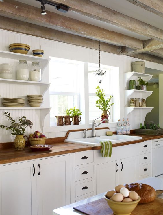a white farmhouse kitchen with butcherblock countertops, open shelves, green touches and reclaimed wooden beams on the ceiling
