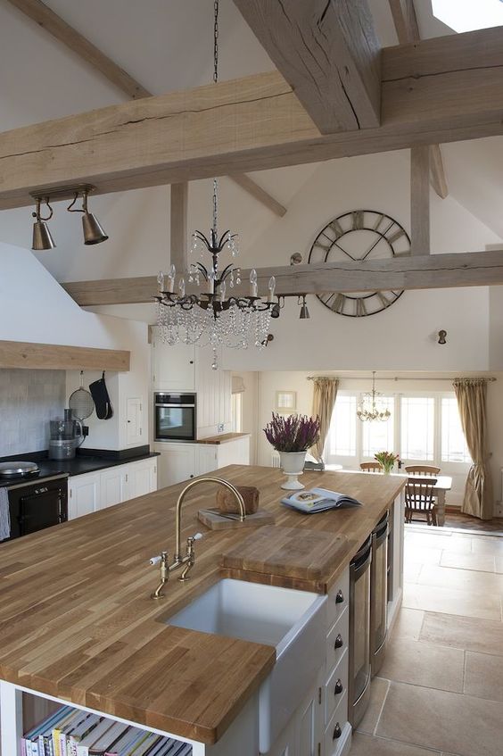 a white kitchen with black and butcherblock countertops,wooden beams, a crystal chandelier and gold fixtures