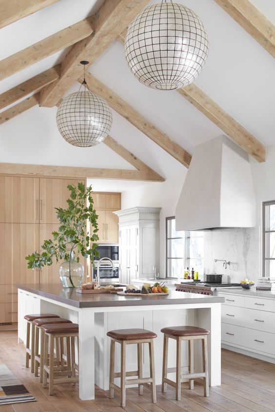 a white light filled kitchen with sleke cabinets, a large hood, wooden beams, sphere lamps, a large kitchen island with an eating space