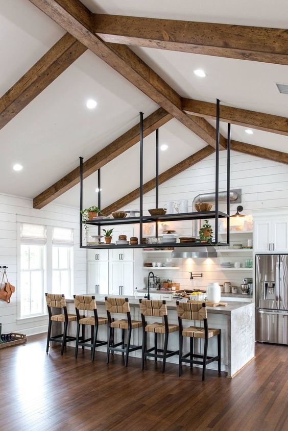 a white modern farmhouse kitchen with planked walls, wooden beams, a large kitchen island, woven chairs and a suspended shelf
