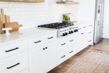 a white modern farmhouse kitchen with shaker cabinets, black fixtures, floating shelves and a hood clad with wood