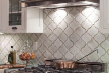 a white retro kitchen with shaker and glass cabinets, a statement black cooker and a hood, a white arabesque tile backsplash and grey stone countertops