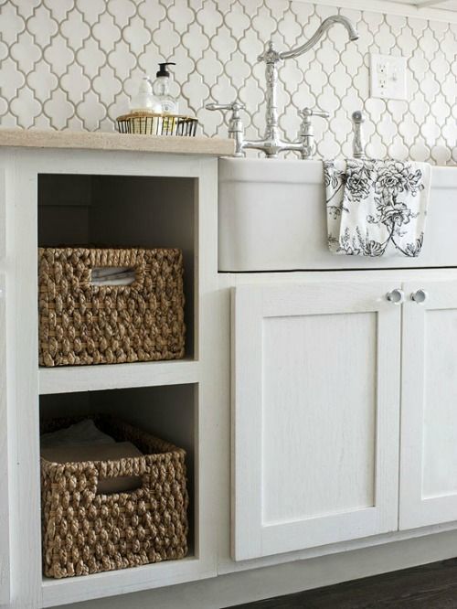 a white vintage kitchen with shaker style cabinets, baskets instead of drawers, a white arabesque tile backsplash and vintage fixtures