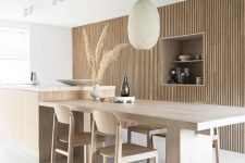 an airy Japandi kitchen with a woode slab wall that hides storage space, a large kitchen island and table here, some lovely chairs