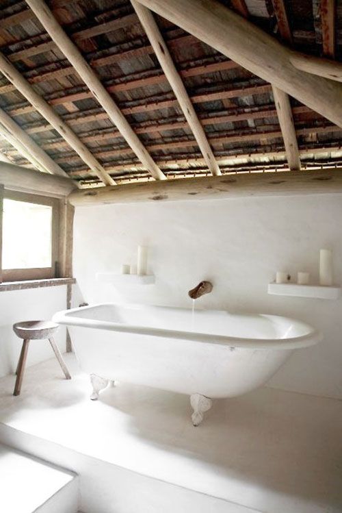 an airy bathroom in white, with a roof with wooden beams, a clawfoot tub, a stool and some shelves with pillar candles is like a spa